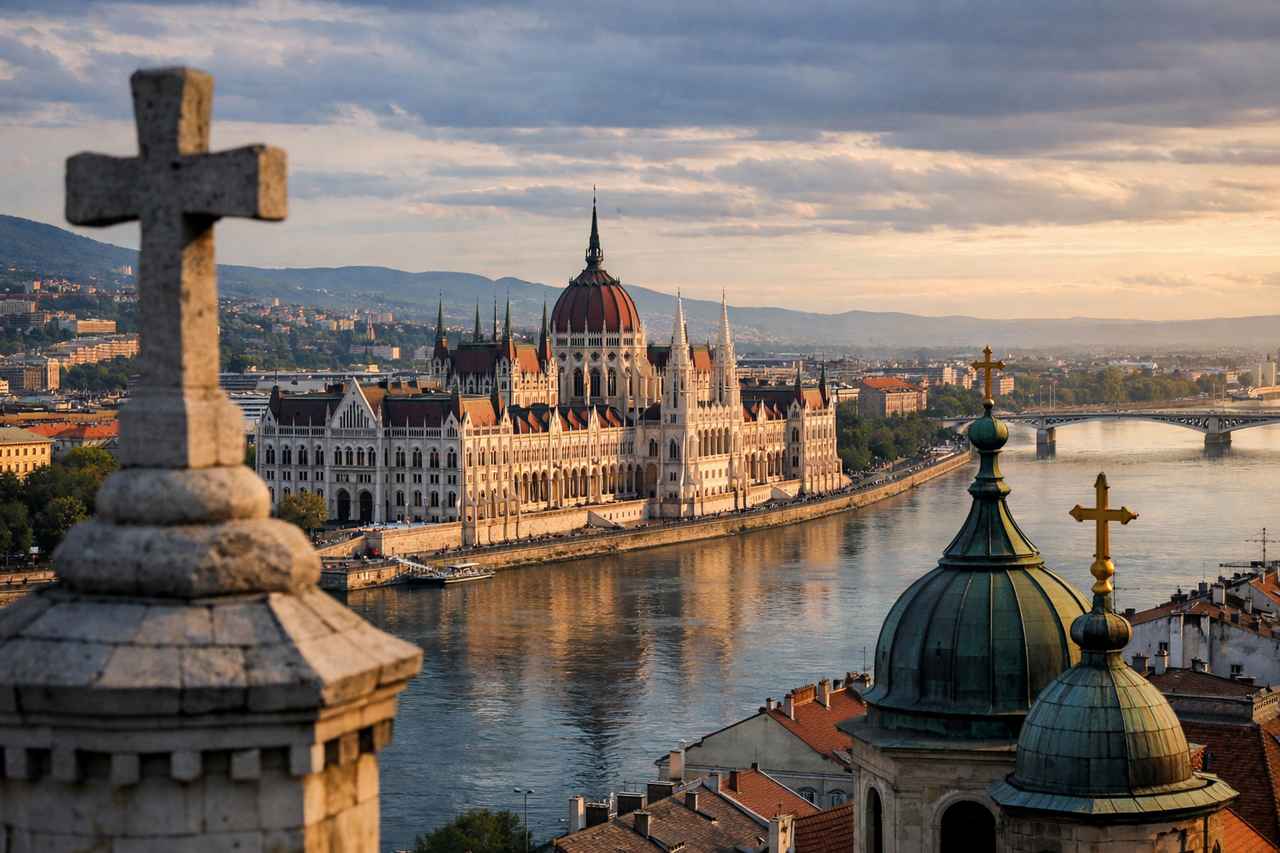 Panoramic view of Budapest, Hungary, with the Parliament building along the Danube River and church domes in the foreground at sunset.