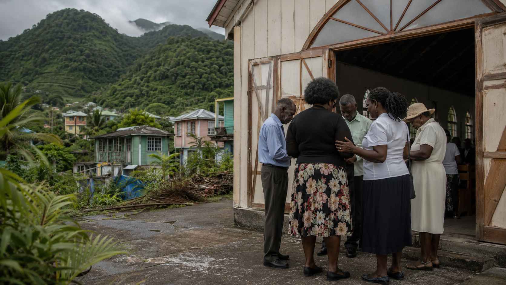 Believers gathered in quiet prayer outside a small church in mountainous Dominica, with village homes, green hills, and a cloudy sky in the background.