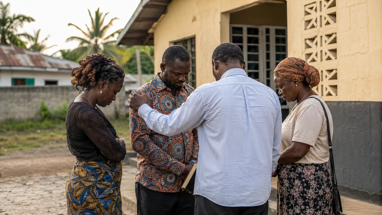 Four adults bow their heads in a prayer circle outside a modest building, with one person resting a supportive hand on another’s shoulder.